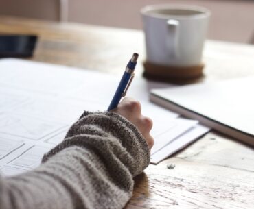 person writing on brown wooden table near white ceramic mug