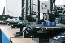 A row of 3D printers creating blue plastic cylinders on a wooden workbench